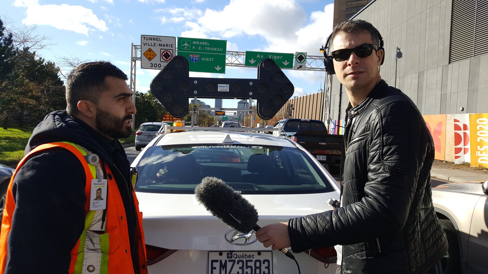 Un homme portant un dossard orange parle dans un micro tenu par un autre homme derrière une voiture portant un panneau lumineux stationnée dans une rue passante. Un homme portant un dossard orange parle dans un micro tenu par un autre homme derrière une voiture portant un panneau lumineux stationnée dans une rue passante.