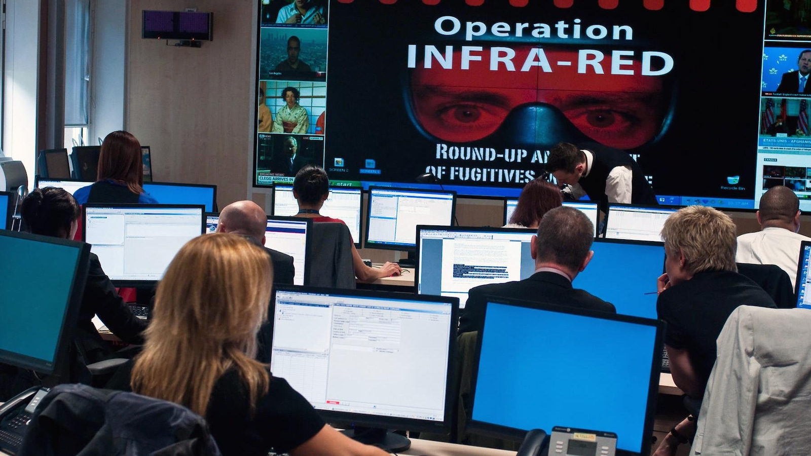 Dans une salle, des personnes travaillent sur des ordinateurs et devant un écran géant portant l'inscription Operation infra-red. Dans une salle, des personnes travaillent sur des ordinateurs et devant un écran géant portant l'inscription Operation infra-red.