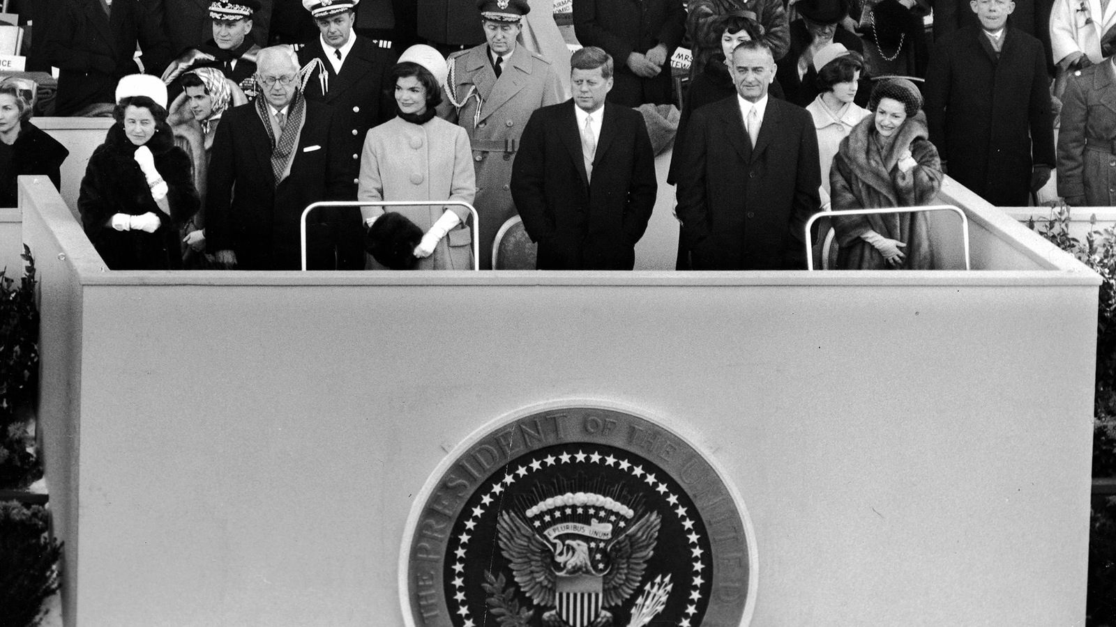 Photo en noir et blanc d'une douzaine de personnes debout sur une plateforme qui porte le sigle du président des États-Unis. Photo en noir et blanc d'une douzaine de personnes debout sur une plateforme qui porte le sigle du président des États-Unis.