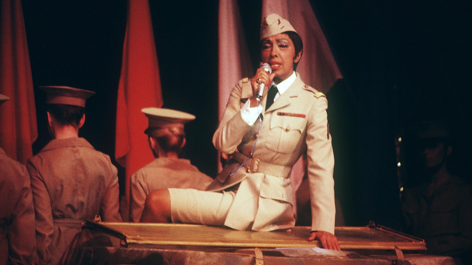 Une femme portant un uniforme militaire chante assise sur un piano. Deux soldats sont derrière-elle et lui font dos. Une femme portant un uniforme militaire chante assise sur un piano. Deux soldats sont derrière-elle et lui font dos.