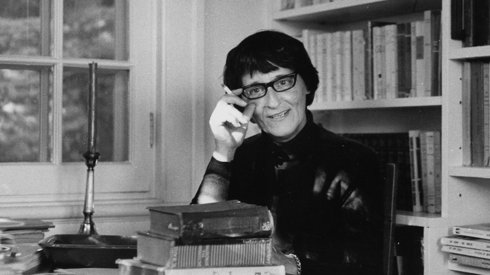 Françoise Loranger, assise à un bureau devant une bibliothèque et une fenêtre, ajuste ses lunettes en fumant une cigarette. Françoise Loranger, assise à un bureau devant une bibliothèque et une fenêtre, ajuste ses lunettes en fumant une cigarette.