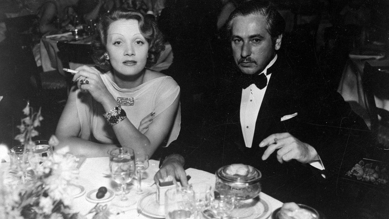 Photo en noir et blanc d'une femme et d'un homme qui regardent la caméra alors qu'ils sont assis à une table ronde dans un restaurant. Les deux ont une cigarette entre leurs doigts. Photo en noir et blanc d'une femme et d'un homme qui regardent la caméra alors qu'ils sont assis à une table ronde dans un restaurant. Les deux ont une cigarette entre leurs doigts.
