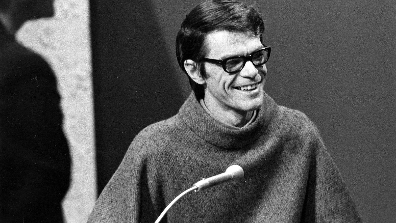 Photo en noir et blanc d'un homme portant des lunettes qui sourit devant un micro. Photo en noir et blanc d'un homme portant des lunettes qui sourit devant un micro.
