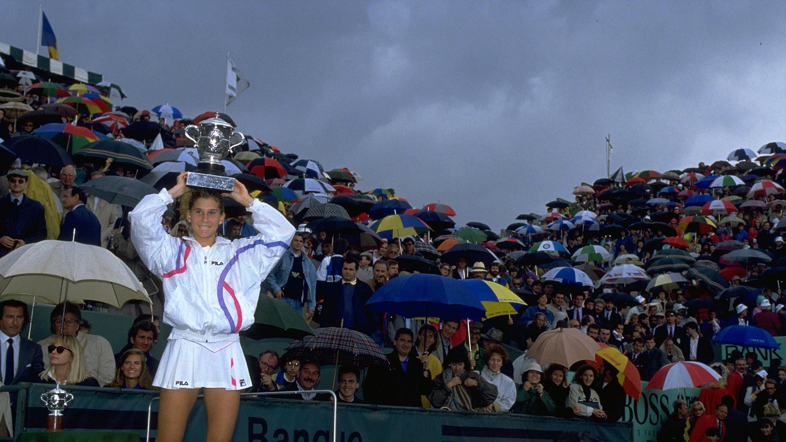 Une joueuse de tennis tient sur sa tête un trophée devant une foule avec des parapluies, assise dans des gradins. Une joueuse de tennis tient sur sa tête un trophée devant une foule avec des parapluies, assise dans des gradins.