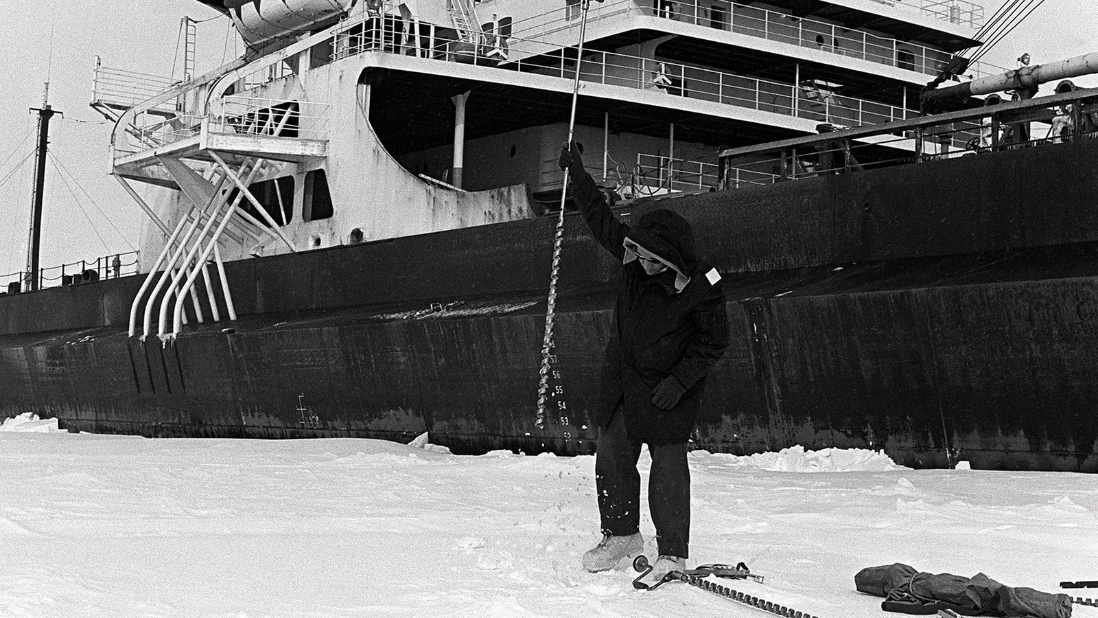 Photo en noir et blanc d'un homme qui lève un pic au-dessus de la glace alors qu'un navire se trouve derrière lui. Photo en noir et blanc d'un homme qui lève un pic au-dessus de la glace alors qu'un navire se trouve derrière lui.