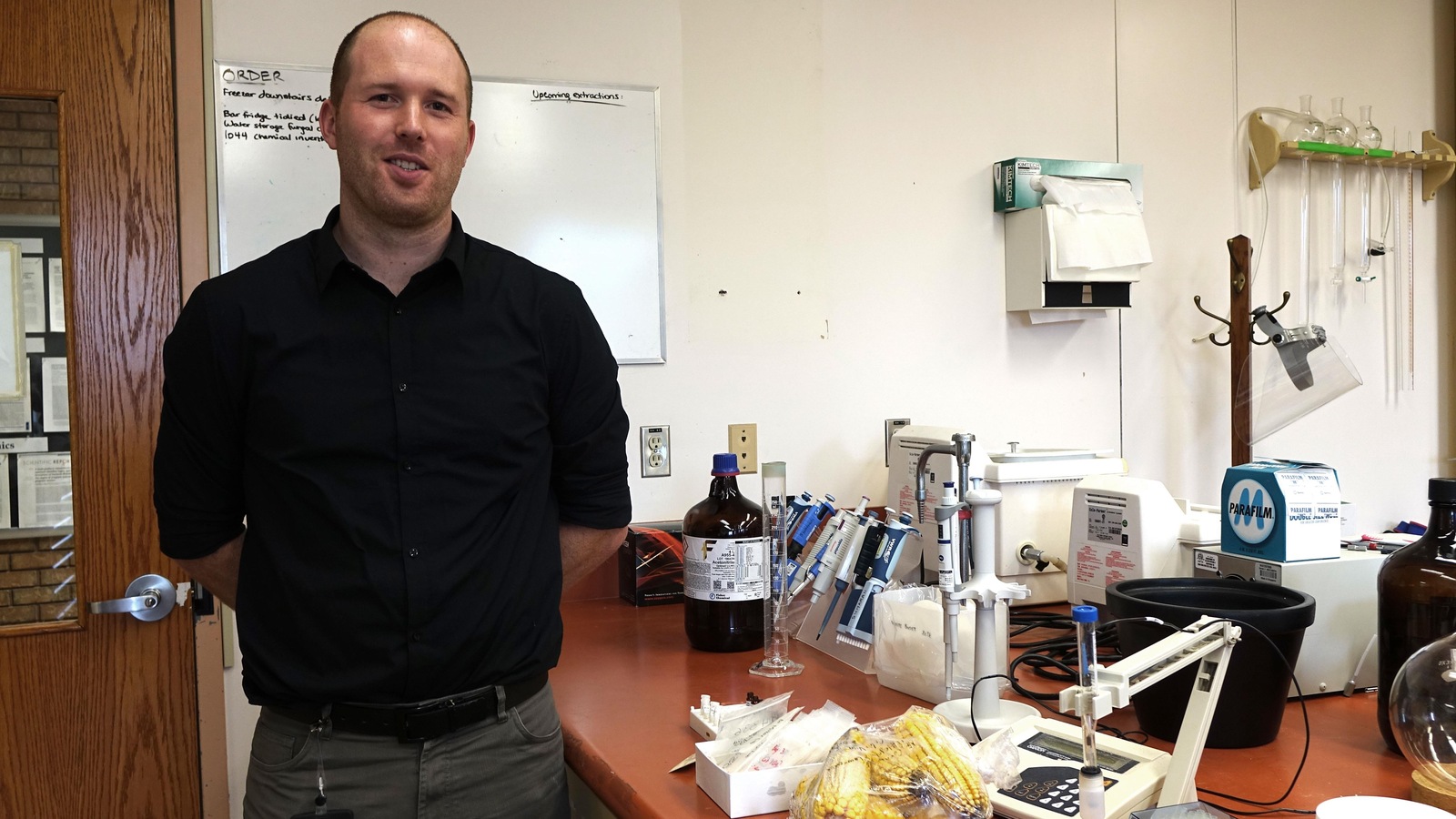Il se tient debout, les bras dans le dos, devant un comptoir où sont déposés des outils scientifiques et un sac de maïs. Il se tient debout, les bras dans le dos, devant un comptoir où sont déposés des outils scientifiques et un sac de maïs.