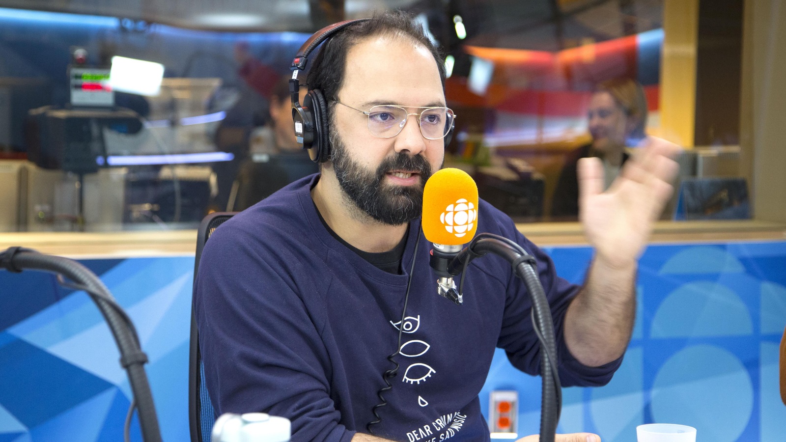 Des écouteurs sur la tête, il est assis derrière un micro et lève le bras gauche en parlant. Des écouteurs sur la tête, il est assis derrière un micro et lève le bras gauche en parlant.
