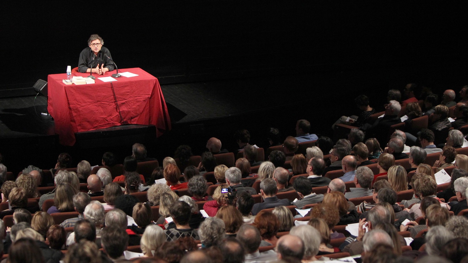 Devant public, il est assis derrière une table avec une nappe rouge. Devant public, il est assis derrière une table avec une nappe rouge.
