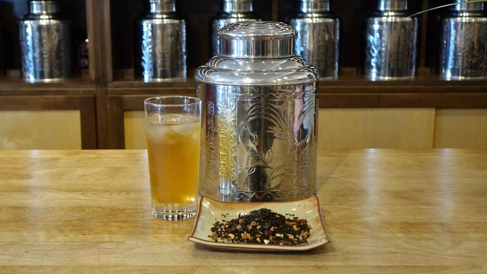 La photo montre un verre de thé glacé, une boîte de thé métallique et une assiette contenant un mélange de thé. La photo montre un verre de thé glacé, une boîte de thé métallique et une assiette contenant un mélange de thé.