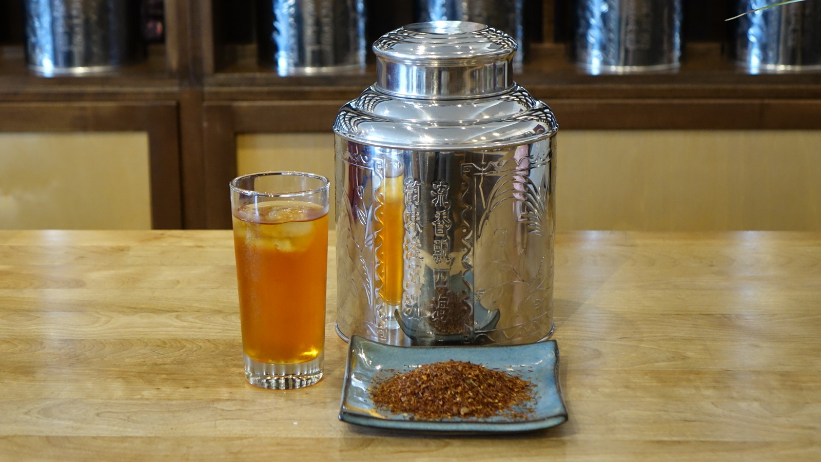La photo montre un verre de thé glacé, une boîte de thé métallique et une assiette contenant un mélange de thé. La photo montre un verre de thé glacé, une boîte de thé métallique et une assiette contenant un mélange de thé.
