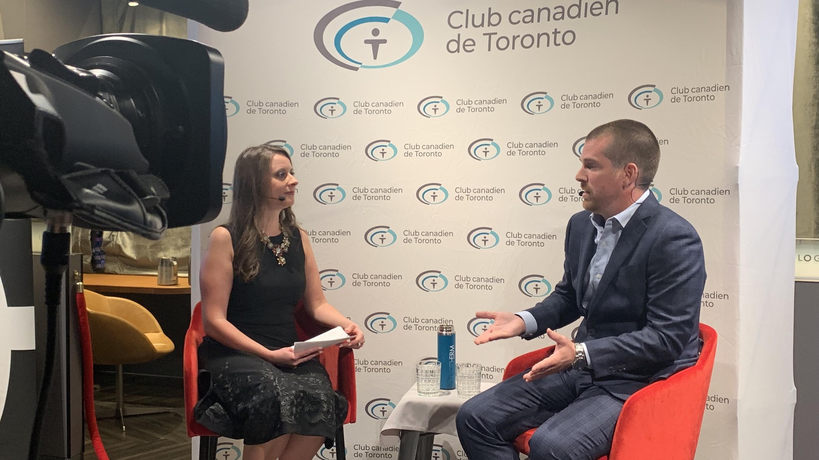 Une femme sur une chaise en discussion avec un homme assis à sa droite. La lentille d'une caméra de télévision apparaît à la gauche. Une femme sur une chaise en discussion avec un homme assis à sa droite. La lentille d'une caméra de télévision apparaît à la gauche.