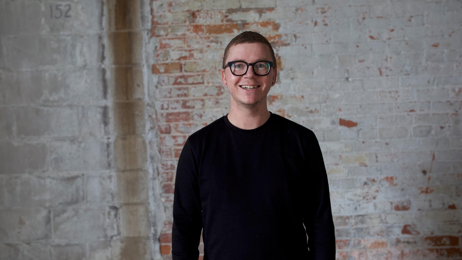 Un homme habillé de noir avec des lunettes à fortes montures pose devant un mur de brique délavé, en souriant. Un homme habillé de noir avec des lunettes à fortes montures pose devant un mur de brique délavé, en souriant.