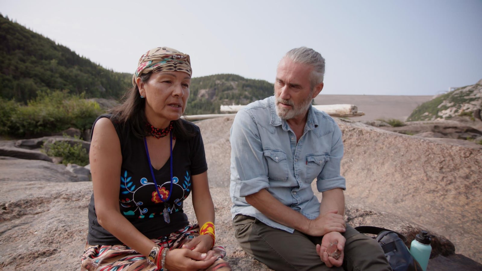 Roy Dupuis et Rita Mestokosho qui discutent assis sur un rocher.