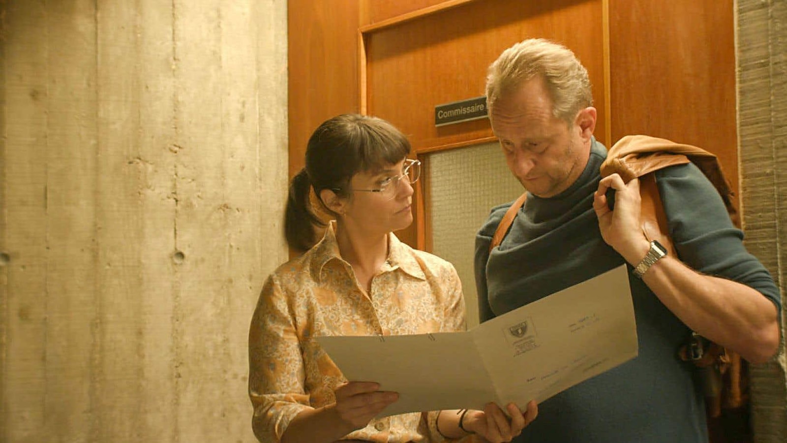 Un homme portant un holster et une veste sur l'épaule regarde un document que lui tend une jeune femme. Un homme portant un holster et une veste sur l'épaule regarde un document que lui tend une jeune femme.