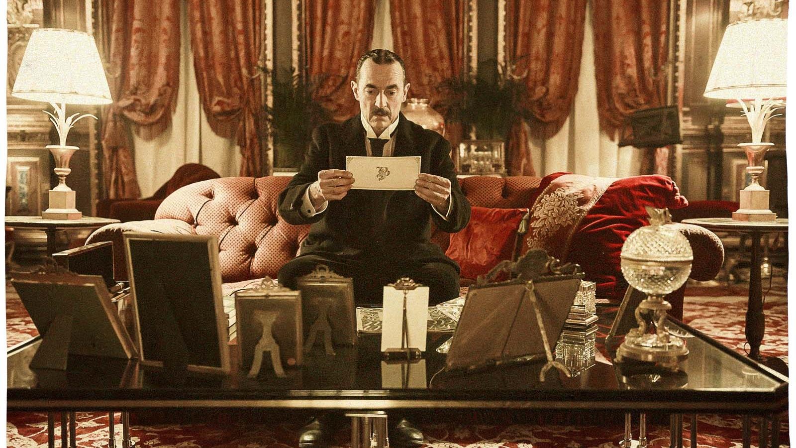 Un homme (Albert Dupontel) en tenue de soirée assis sur un canapé dans une pièce luxueuse lit un carton. Un homme (Albert Dupontel) en tenue de soirée assis sur un canapé dans une pièce luxueuse lit un carton.