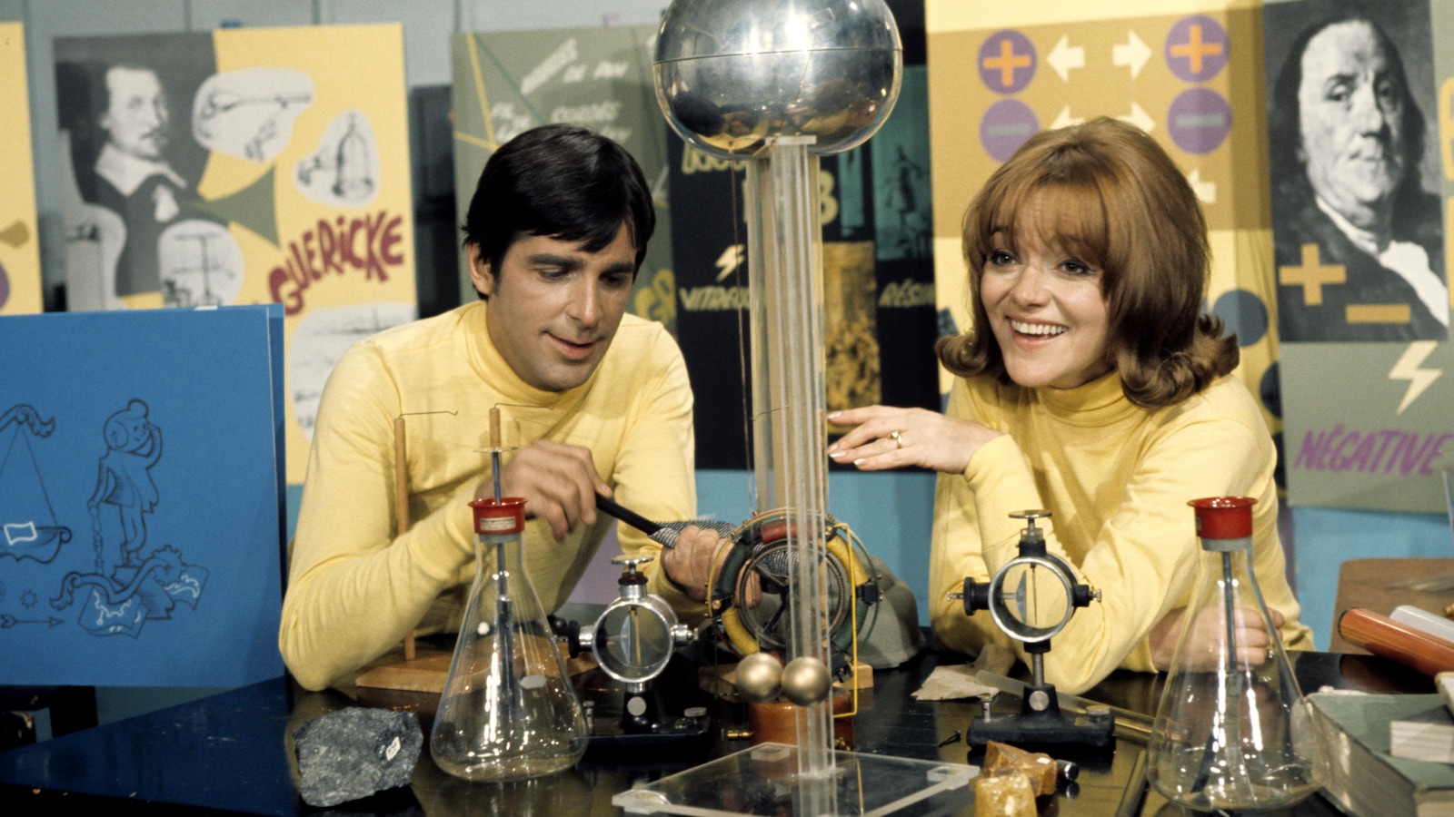 Photo d'archives de 1969 : Dans un décor de laboratoire, les animateurs Jean Besré et Lise Lasalle, font une expérience scientifique avec des objets disposés sur la table devant eux 