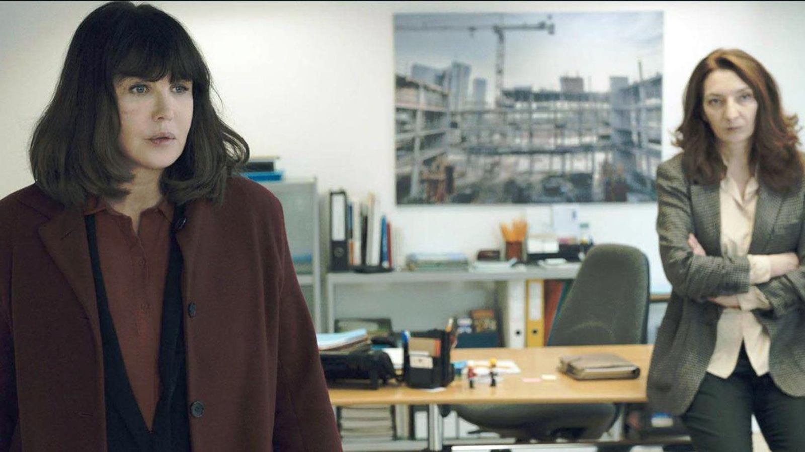 Une femme en manteau bourgogne en avant plan, une autre en tailleur gris, en arrière-plan, dans un bureau. Une femme en manteau bourgogne en avant plan, une autre en tailleur gris, en arrière-plan, dans un bureau.