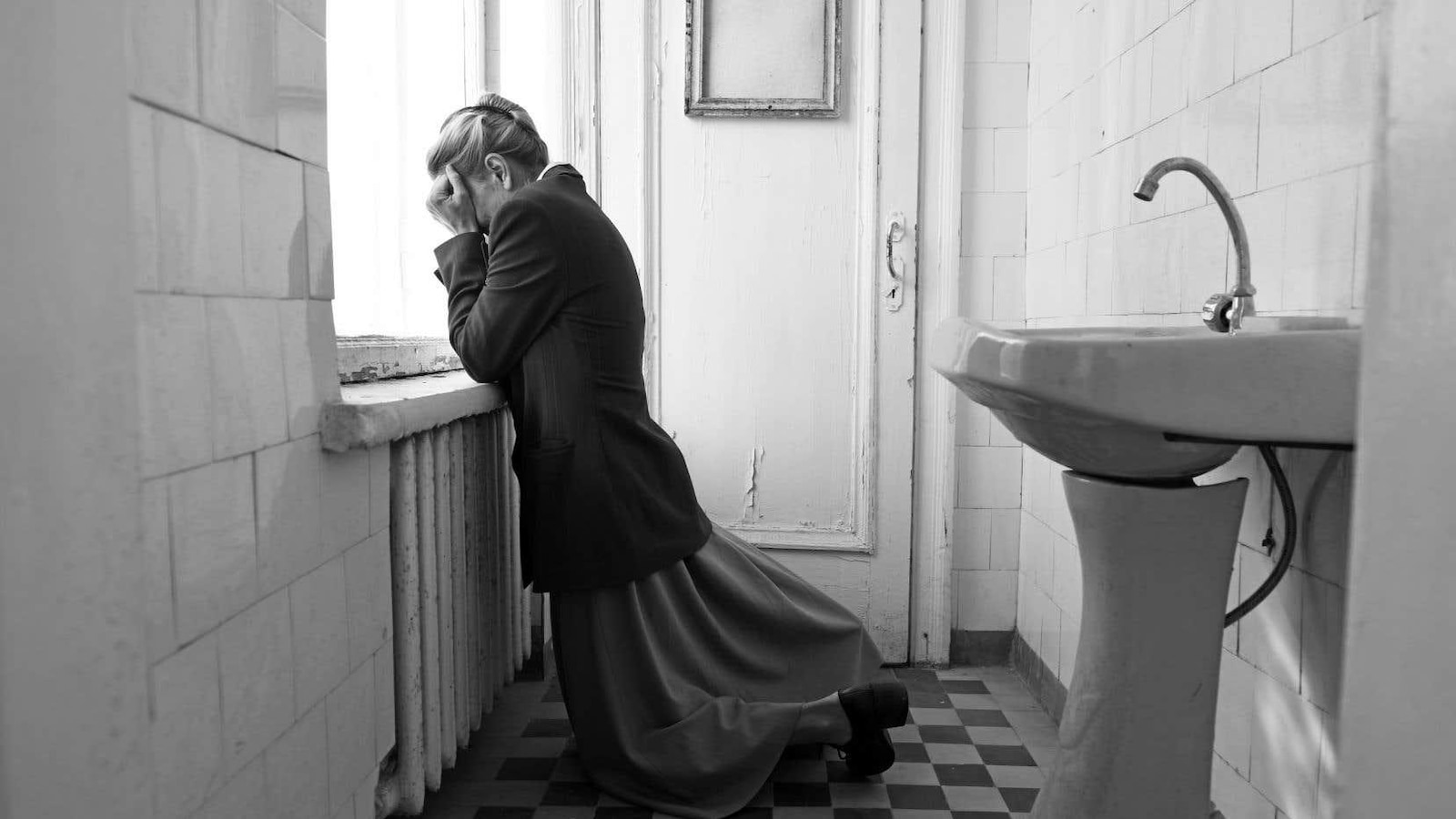 En noir et blanc, une femme à genoux appuyée contre un rebord de fenêtre, le visage entre les mains.
