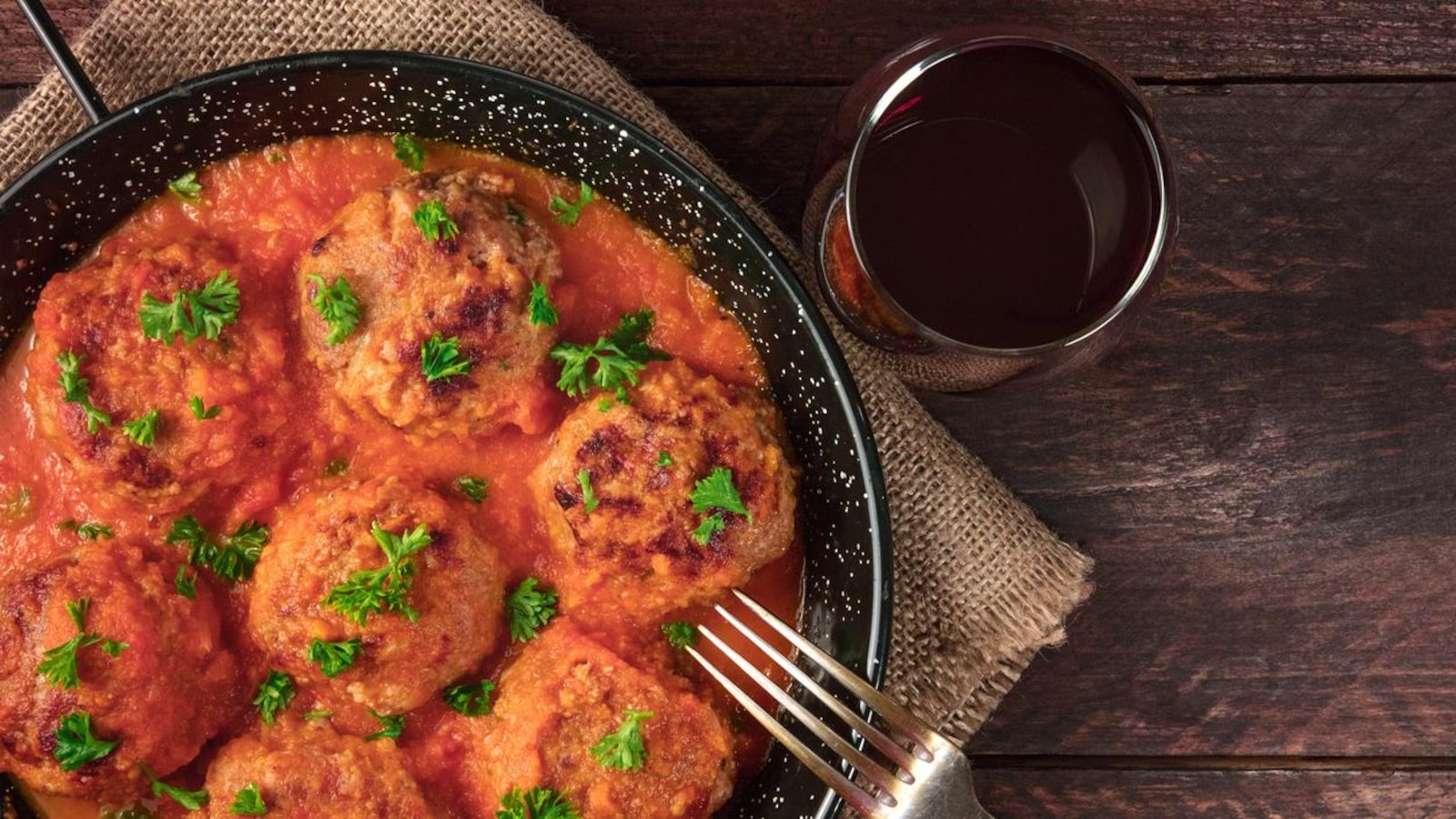 An overhead photo of meatballs in tomato sauce, shot from above in the skillet on dark rustic textures, with a place for text, a fork, and a glass of red wine An overhead photo of meatballs in tomato sauce, shot from above in the skillet on dark rustic textures, with a place for text, a fork, and a glass of red wine