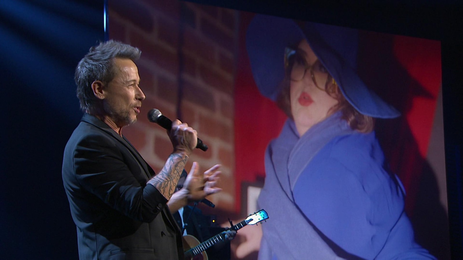 Stéphane Rousseau chante au micro, en arrière plan, Christine Morency est habillée en bleu.