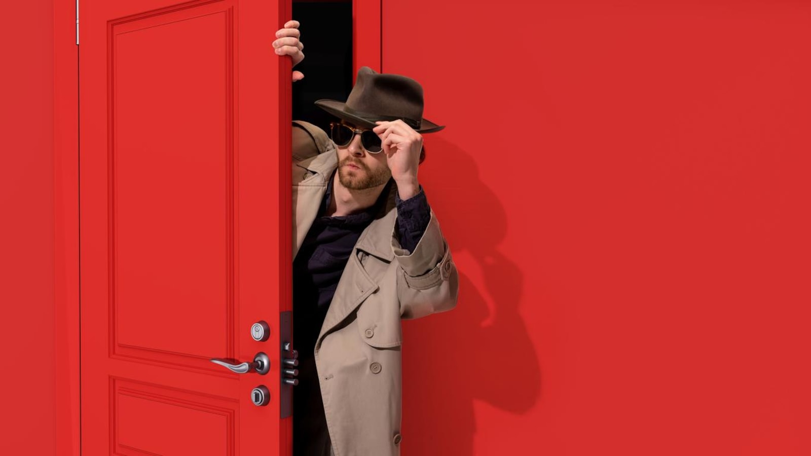 Un homme qui porte un chapeau, des lunettes fumées et un trench dans l'entrebâillement d'une porte.