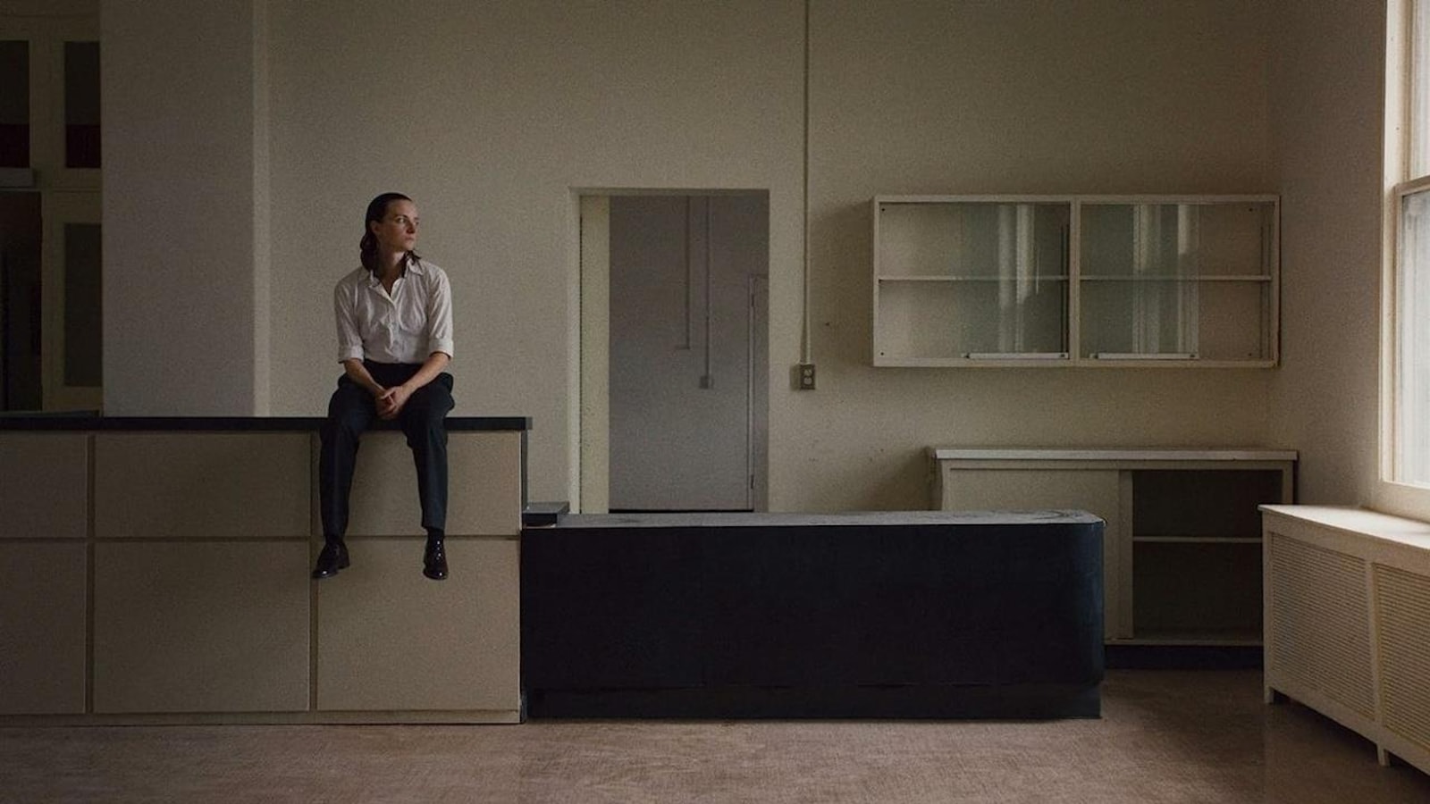 Une femme (Larissa Corriveau) assise sur le rebord d'un comptoir dans une pièce vide.