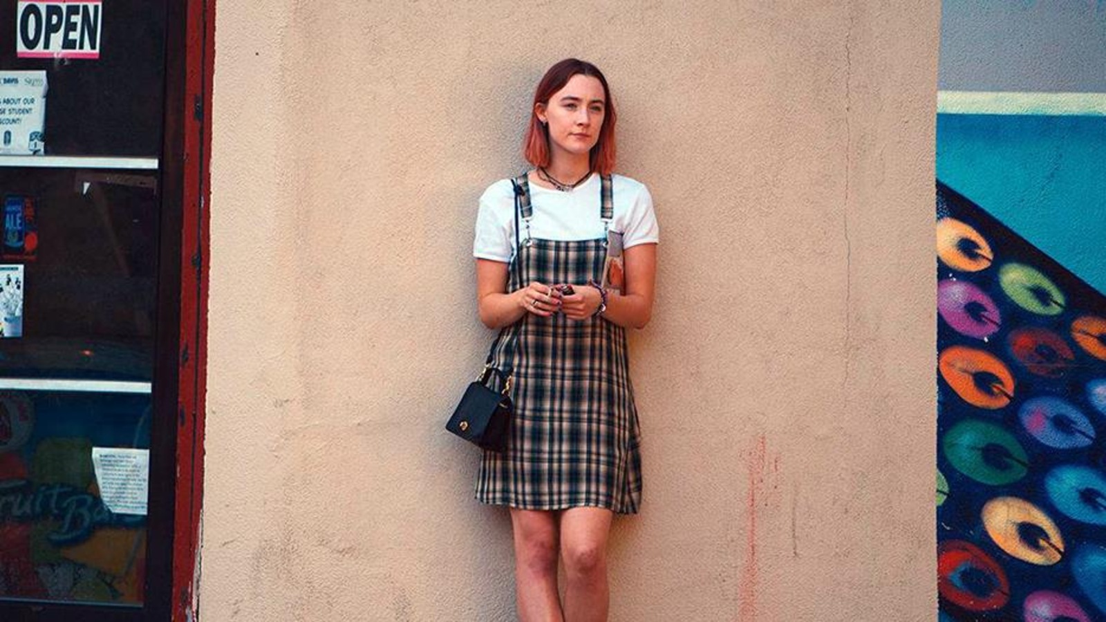 Une jeune fille (Saoirse Ronan) en robe à carreaux, adossée contre un mur beige, à l'extérieur. Une jeune fille (Saoirse Ronan) en robe à carreaux, adossée contre un mur beige, à l'extérieur.