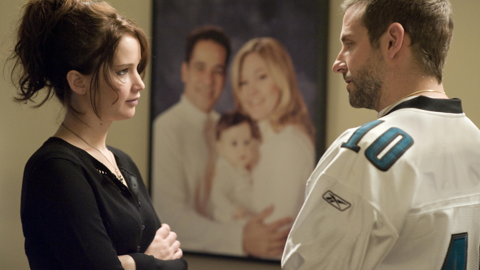 Une femme (Jennifer Lawrence) et un homme (Bradley Cooper) face à face devant un portrait de famille. Une femme (Jennifer Lawrence) et un homme (Bradley Cooper) face à face devant un portrait de famille.