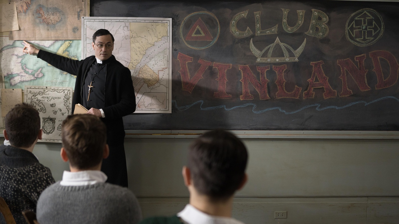 Un prêtre (Sébastien Ricard) pointe un tableau affichant des cartes et la mention Club Vinland à des élèves assis devant lui. Un prêtre (Sébastien Ricard) pointe un tableau affichant des cartes et la mention Club Vinland à des élèves assis devant lui.