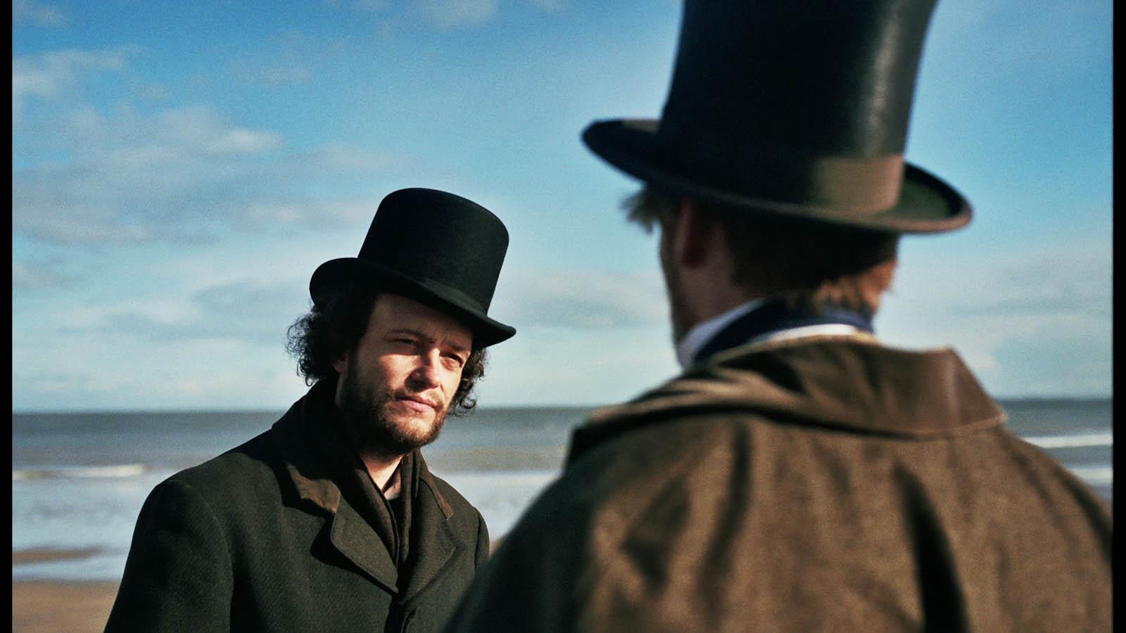On a beach, two men wearing top hats face each other.