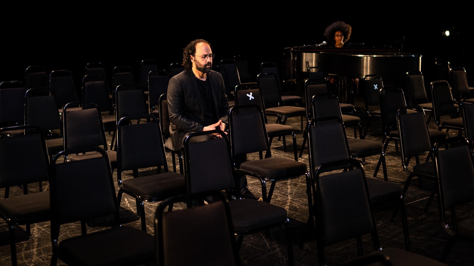 Un homme assis sur une chaise dans une salle remplie de chaises. Il y a aussi un homme au piano.