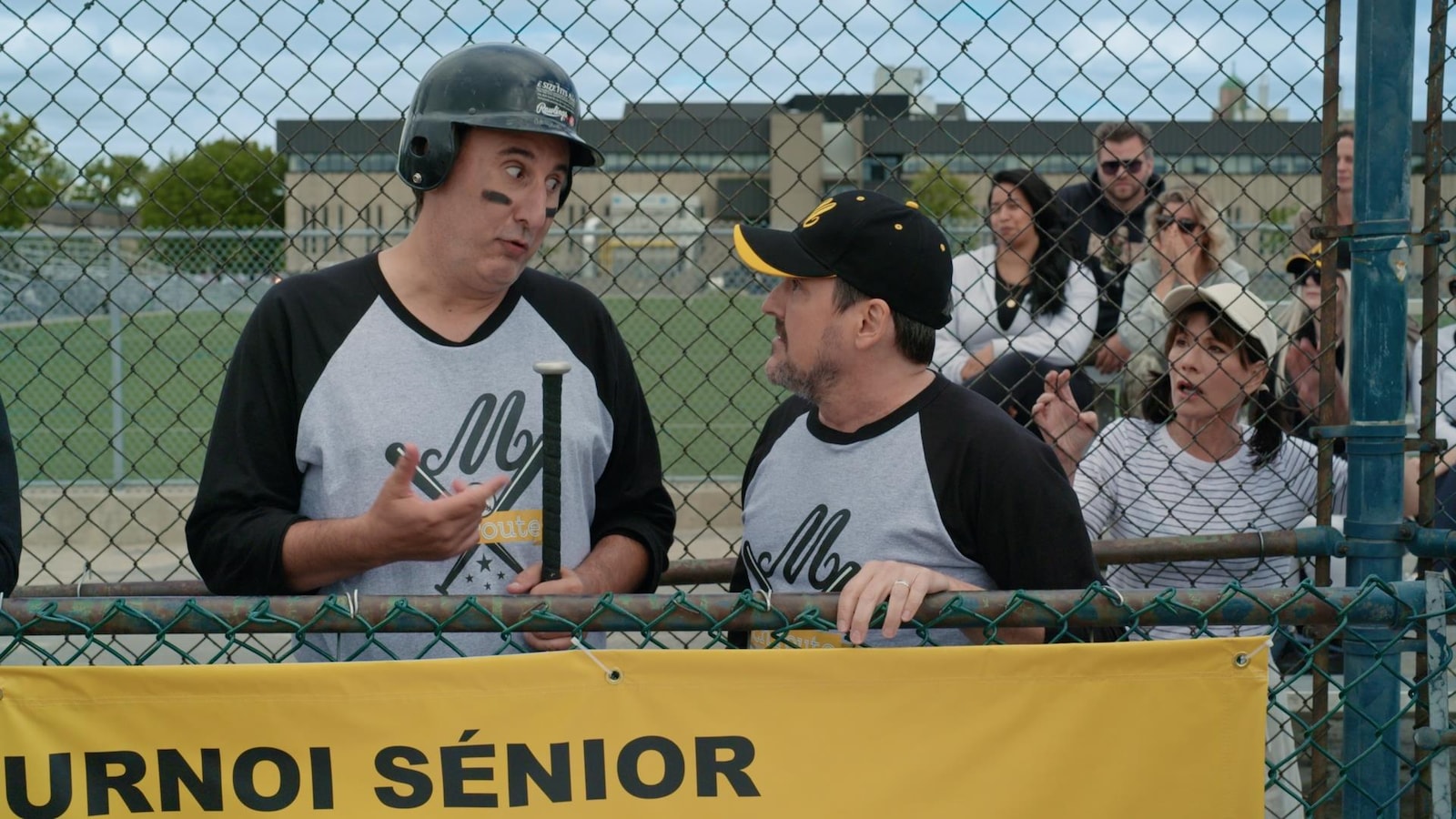 Martin parle avec Guy sur une terrain de baseball, alors que Sylvie les observe à travers une clôture.