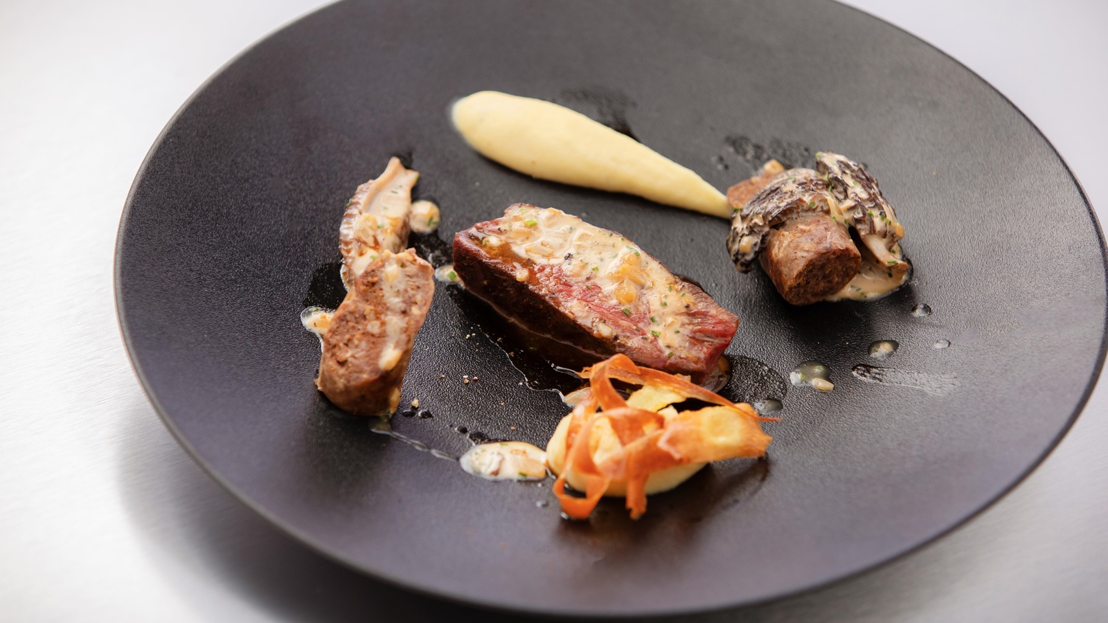 Dans une assiette noire, des morceaux disposés de lamelles de bison, de la purée et de la saucisse. Dans une assiette noire, des morceaux disposés de lamelles de bison, de la purée et de la saucisse.