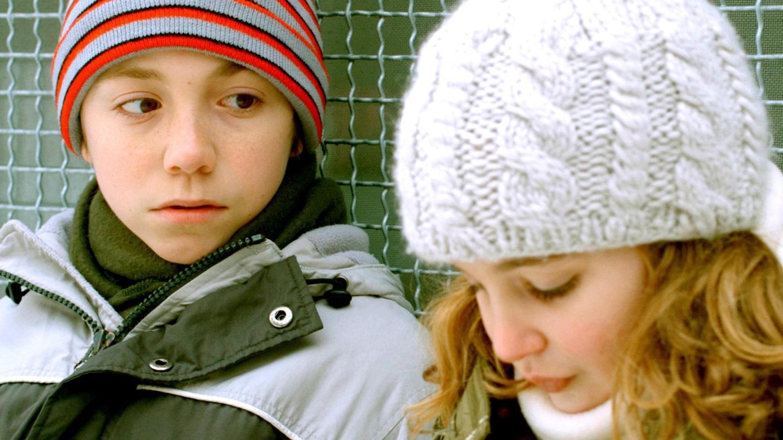 Devant un grillage, portant des habits de neige, un petit garçon regarde une petite fille.