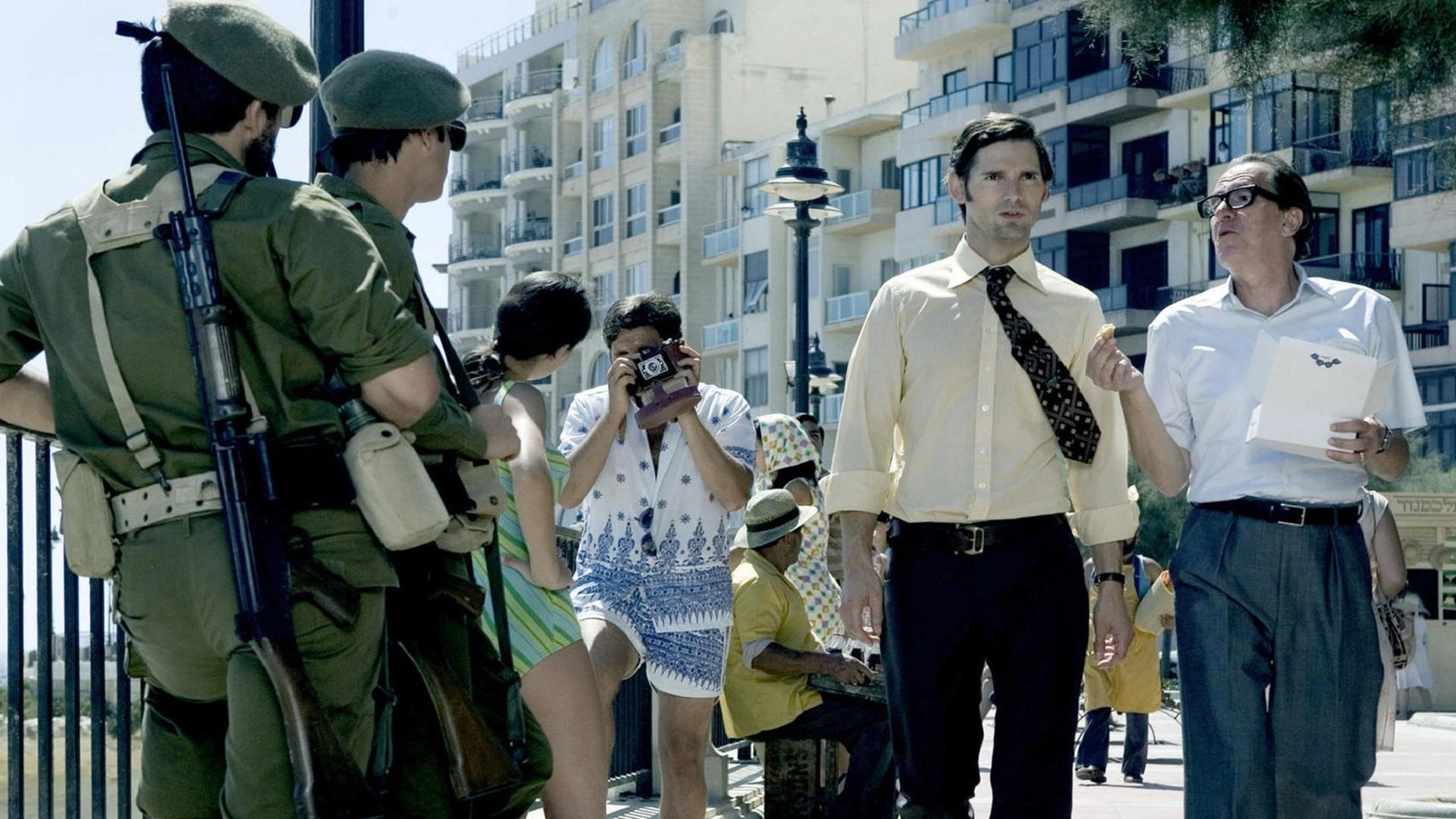 Dans une rue animée, deux hommes en costumes marchent devant des soldats armés. Dans une rue animée, deux hommes en costumes marchent devant des soldats armés.