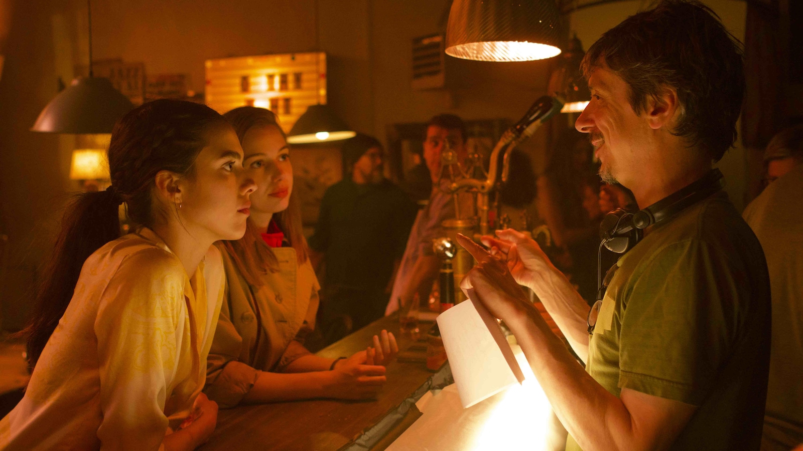 Dans un bar sous une lumière rouge, un homme derrière le bar portant des écouteurs autour du cou (Philippe Falardeau) parle à deux jeunes femmes accoudées sur le bar. Dans un bar sous une lumière rouge, un homme derrière le bar portant des écouteurs autour du cou (Philippe Falardeau) parle à deux jeunes femmes accoudées sur le bar.