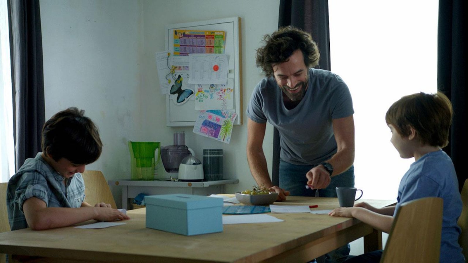 Un homme (Romain Duris) rit, debout devant une table où sont assis deux petits garçons. Un homme (Romain Duris) rit, debout devant une table où sont assis deux petits garçons.