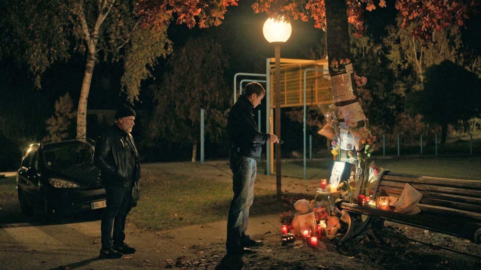 La nuit, deux hommes se recueillent devant un autel spontané près d'un banc de parc.