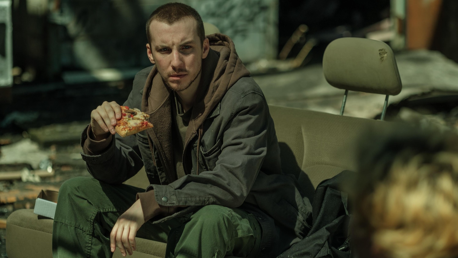 Assis sur une vieille banquette de voiture dans un terrain vague, Jessy (Pier-Luc Funk) tient une pointe de pizza dans la main.