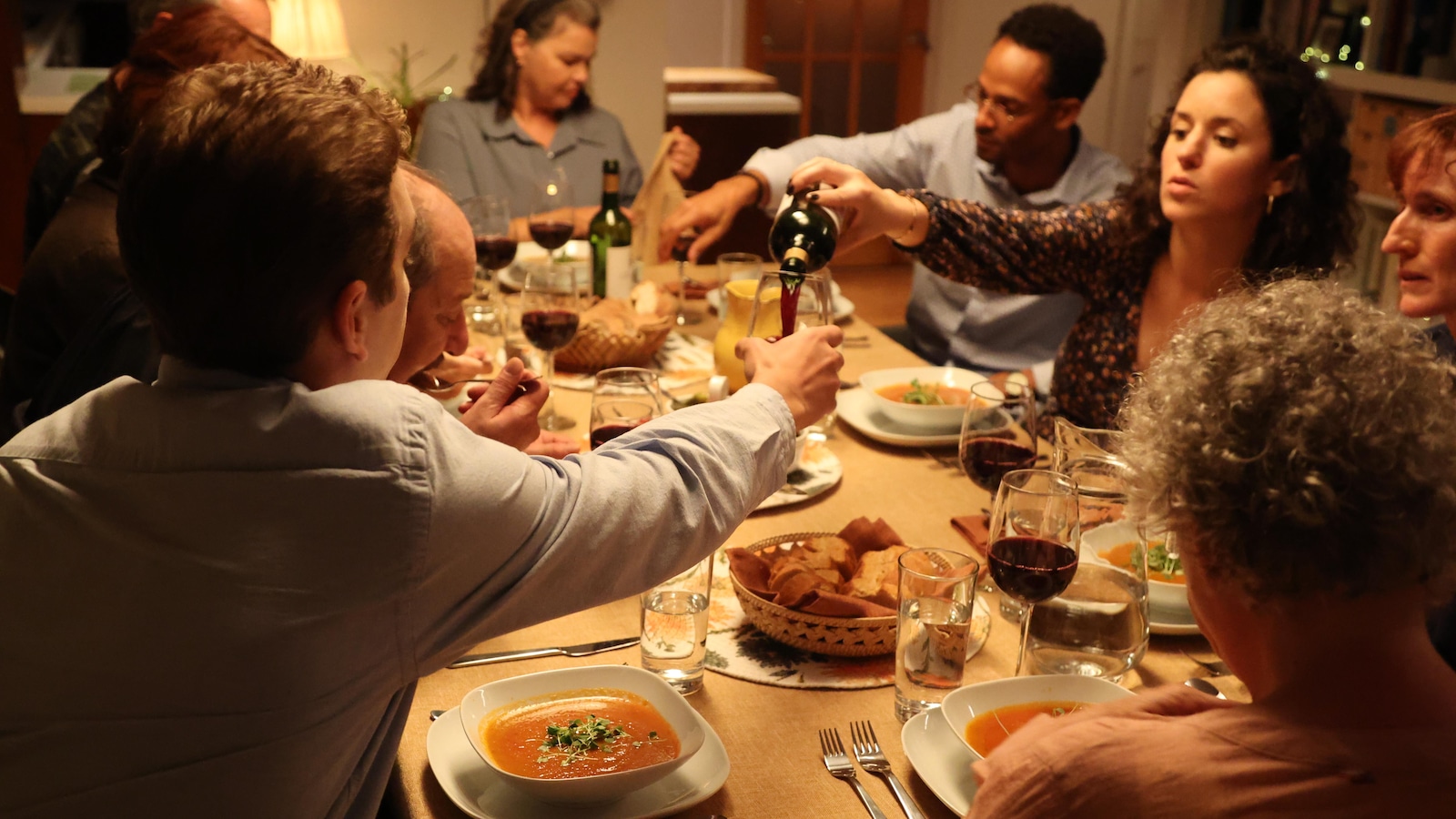 Several people share a meal at a table and one person serves red wine to one of the guests. 
