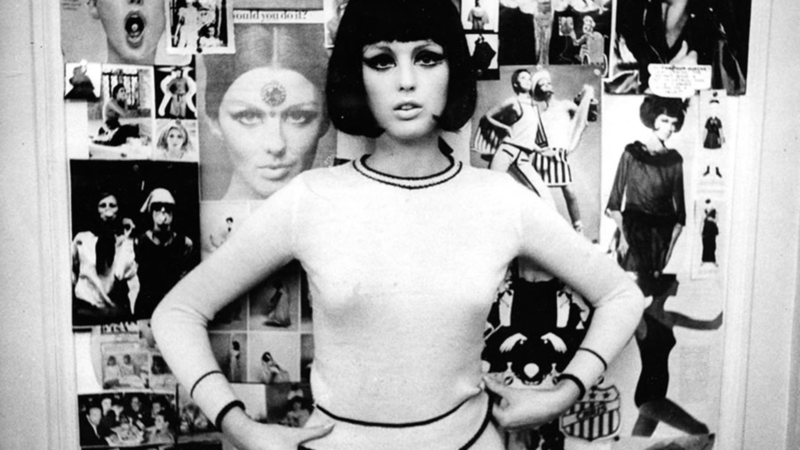 En noir et blanc, une jeune femme brune, habillée en blanc, devant un mur recouvert de photos de mode. En noir et blanc, une jeune femme brune, habillée en blanc, devant un mur recouvert de photos de mode.