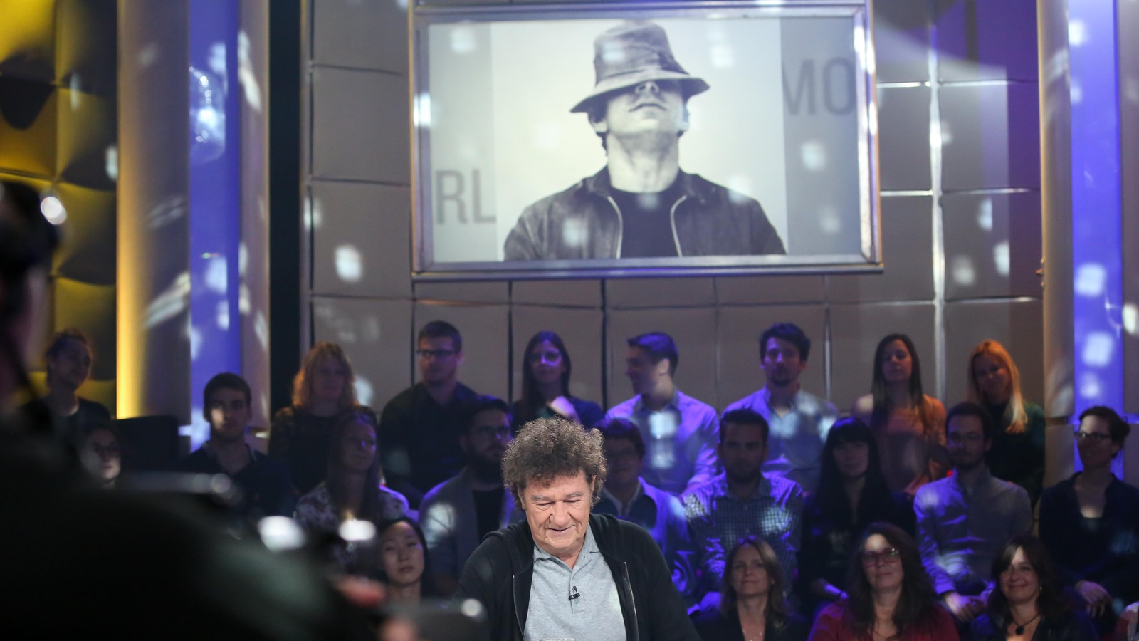 Robert Charlebois est à la table des invités. Une photo de Réjean Ducharme est projetée sur un grand écran. Robert Charlebois est à la table des invités. Une photo de Réjean Ducharme est projetée sur un grand écran.