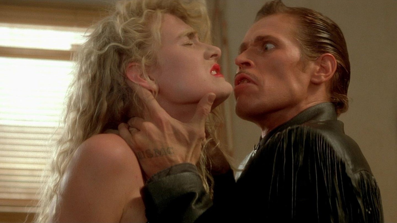 Un homme portant le cheveu gominé et une veste à franges (Willem Dafoe) tient une femme blonde (Laura Dern) par la gorge. Un homme portant le cheveu gominé et une veste à franges (Willem Dafoe) tient une femme blonde (Laura Dern) par la gorge.