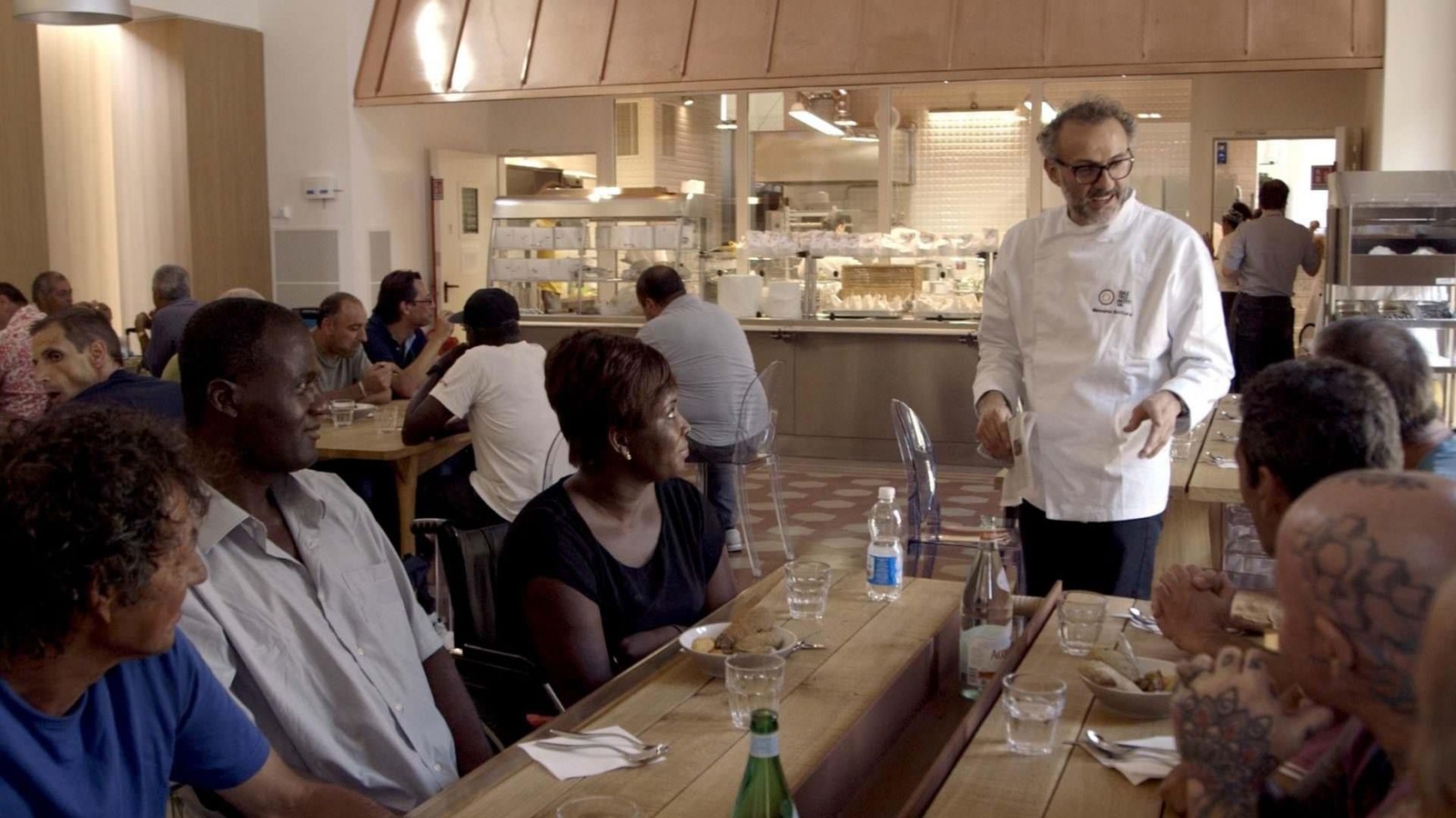 Un cuisinier en veste blanche devant une tablée de gens assis qui le regardent. Un cuisinier en veste blanche devant une tablée de gens assis qui le regardent.