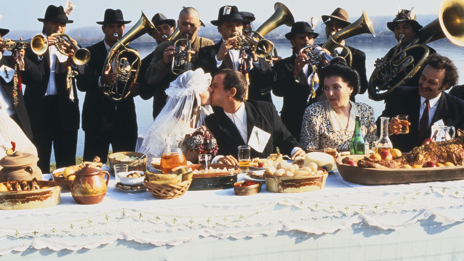 Un couple de mariés assis à table s'embrasse devant une fanfare. Un couple de mariés assis à table s'embrasse devant une fanfare.