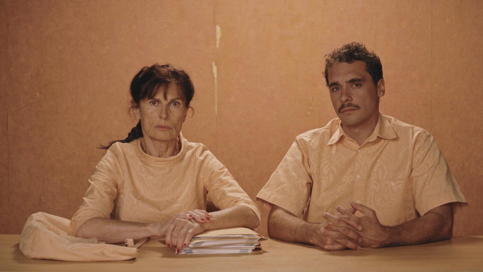 Un homme et une femme, habillés en orange pâle, assis, devant un mur orange pâle. Un homme et une femme, habillés en orange pâle, assis, devant un mur orange pâle.