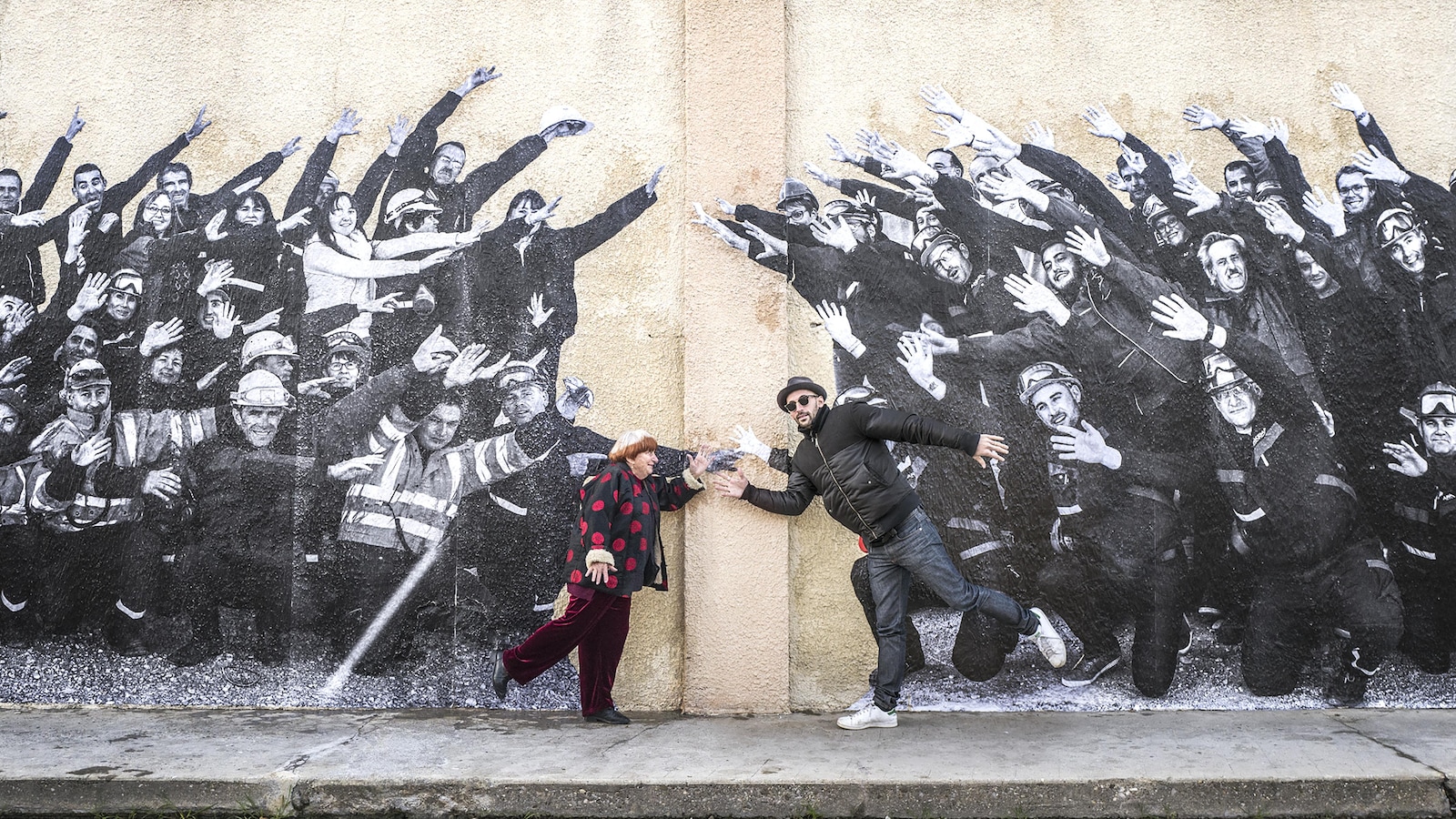 La cinéaste Agnès Varda et le photographe JR posent en s'amusant devant un mur décoré de photos en noir et blanc de villageois et villageoises.