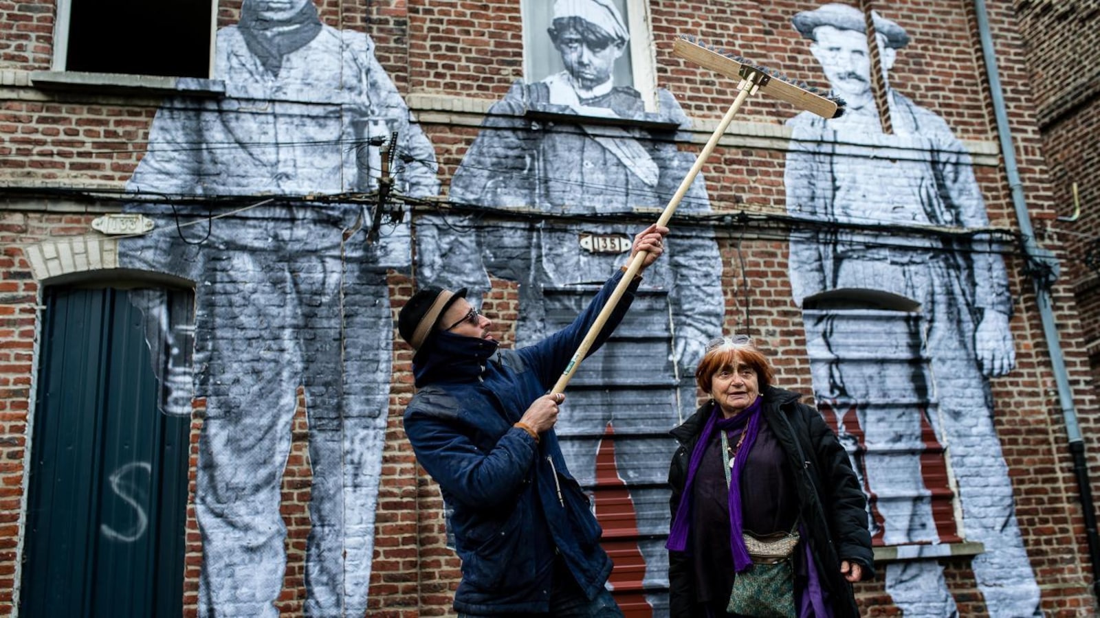 Un homme avec un grand balai colle des photos sur un mur en briques, une femme à ses côtés regarde. Un homme avec un grand balai colle des photos sur un mur en briques, une femme à ses côtés regarde.