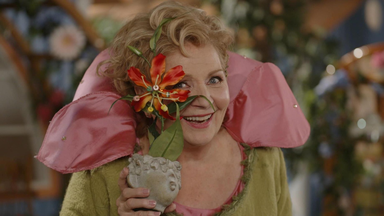 Fée Sylvestre (France Castel) tient un pot de fleur vis-à-vis son visage et regarde à travers les pétales.