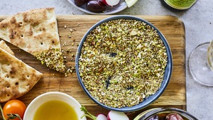 Un bol de dukka aux pistaches et aux amandes sur une planche en bois avec des légumes et du pain autour. 
